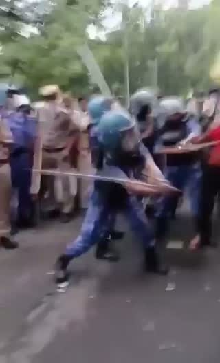 Still from the clip: a helmeted paramilitary officer swinging a lathi on a street, with other personnel and protesters around.